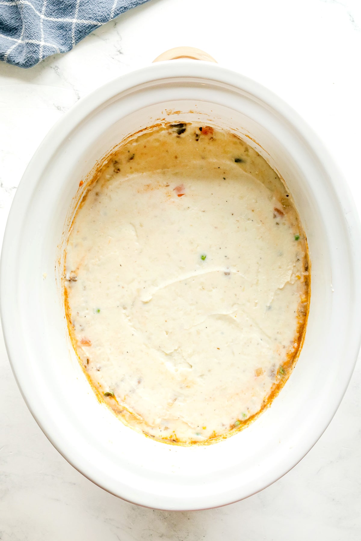 A white slow cooker with remnants of mashed potato and a few visible vegetable pieces, set on a white surface with a blue cloth partially visible in the corner. This is one of the steps in cooking Crockpot Shepherd's Pie