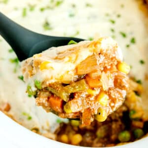 A serving spoon holds a portion of Crockpot Shepherd's Pie with mashed potatoes, mixed vegetables, and ground meat filling.