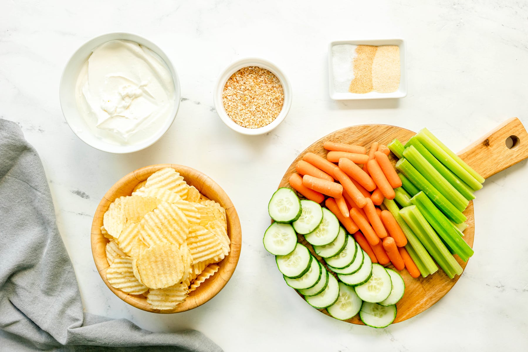 Ingredients for French Onion Dip include: sour cream, dried minced onion, onion powder, garlic powder, and salt. A bowl of A bowl of potato chips, sliced cucumbers, carrots, and celery can also be seen on wooden board.