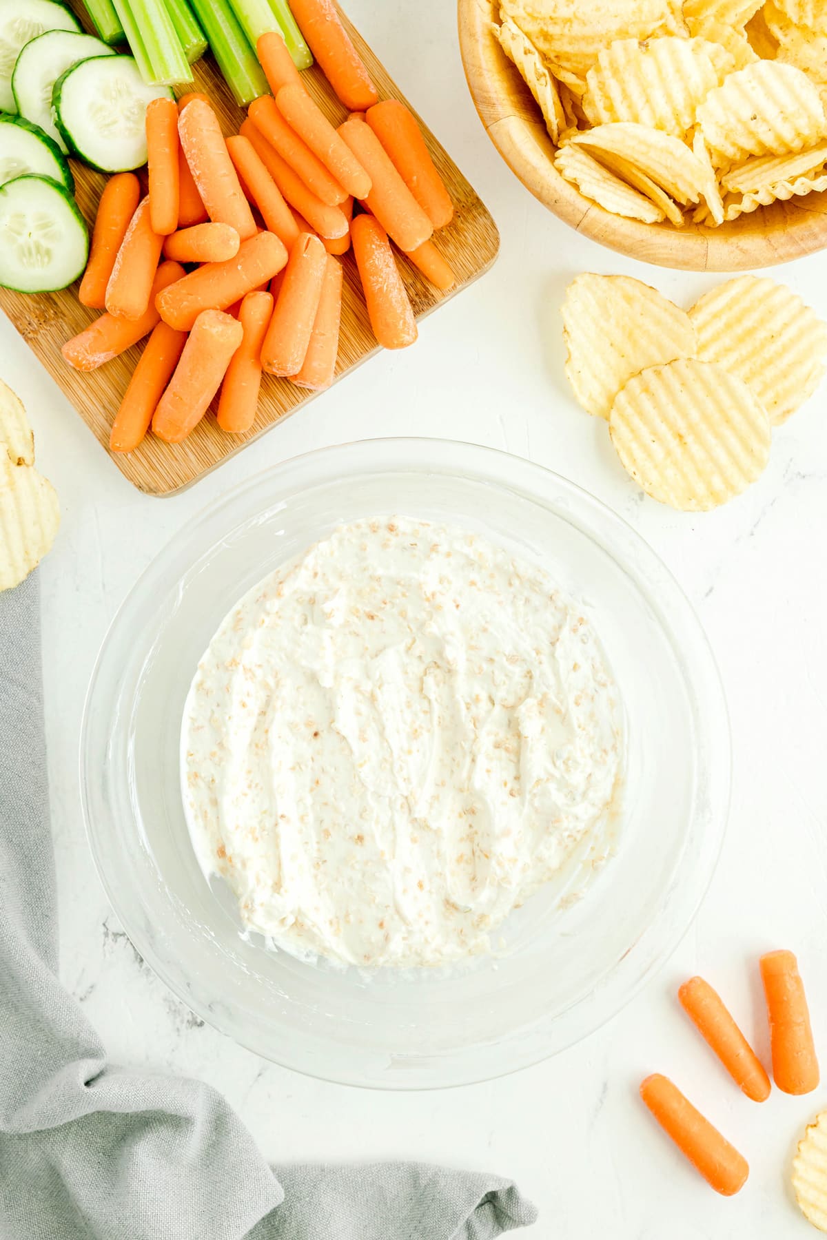 Clear glass bowl of French Onion Dip on a white surface, surrounded by potato chips, baby carrots, celery, cucumber slices, and a gray napkin.