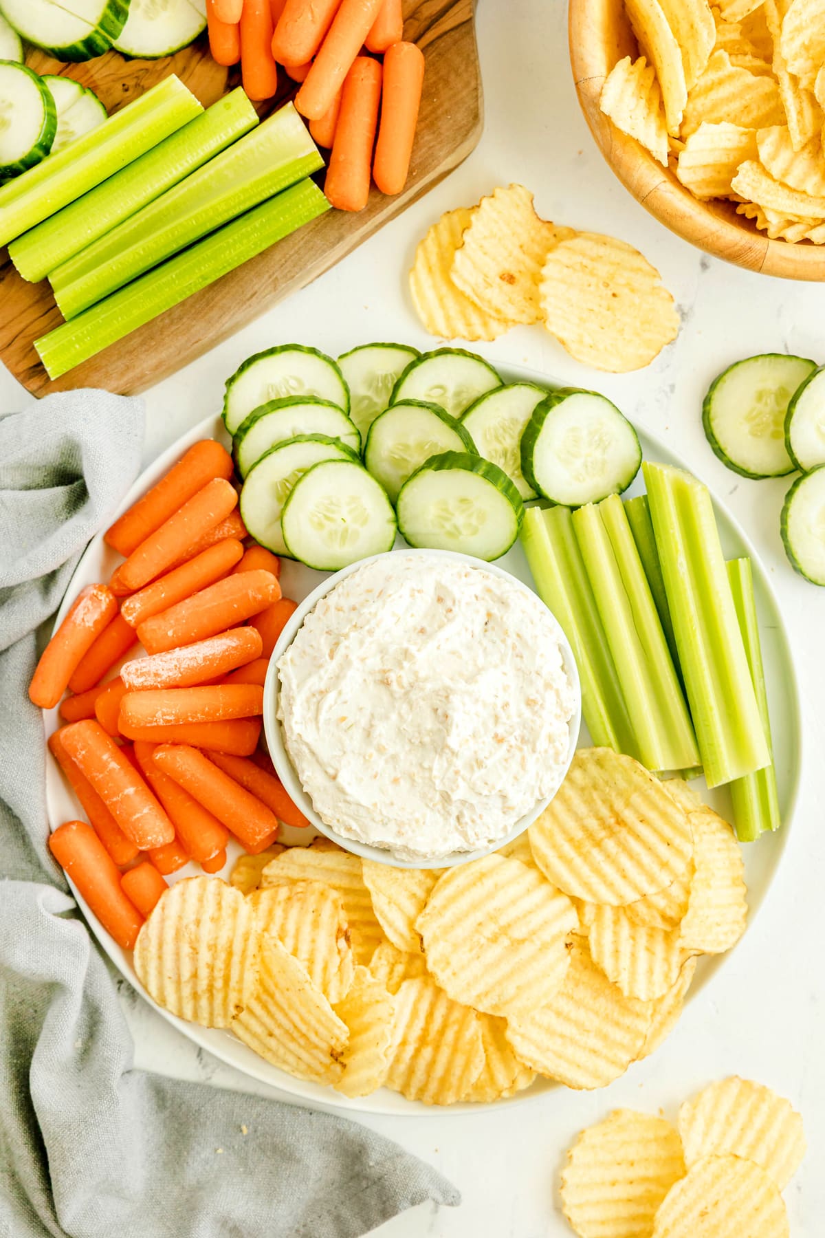 Plate with potato chips, cucumber slices, celery sticks, baby carrots, and a bowl of French Onion Dip in the center. Additional chips and veggies are visible nearby.