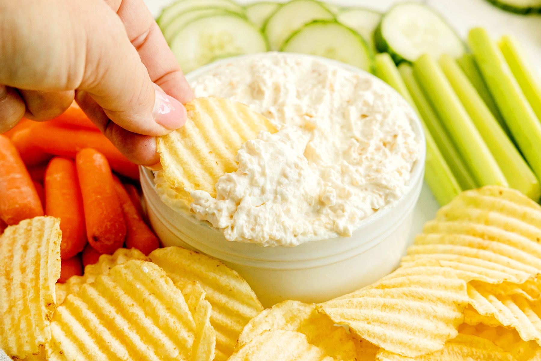 Hand dipping a ridged potato chip into a bowl of creamy French Onion Dip, surrounded by sliced cucumbers, celery sticks, carrots, and more potato chips.