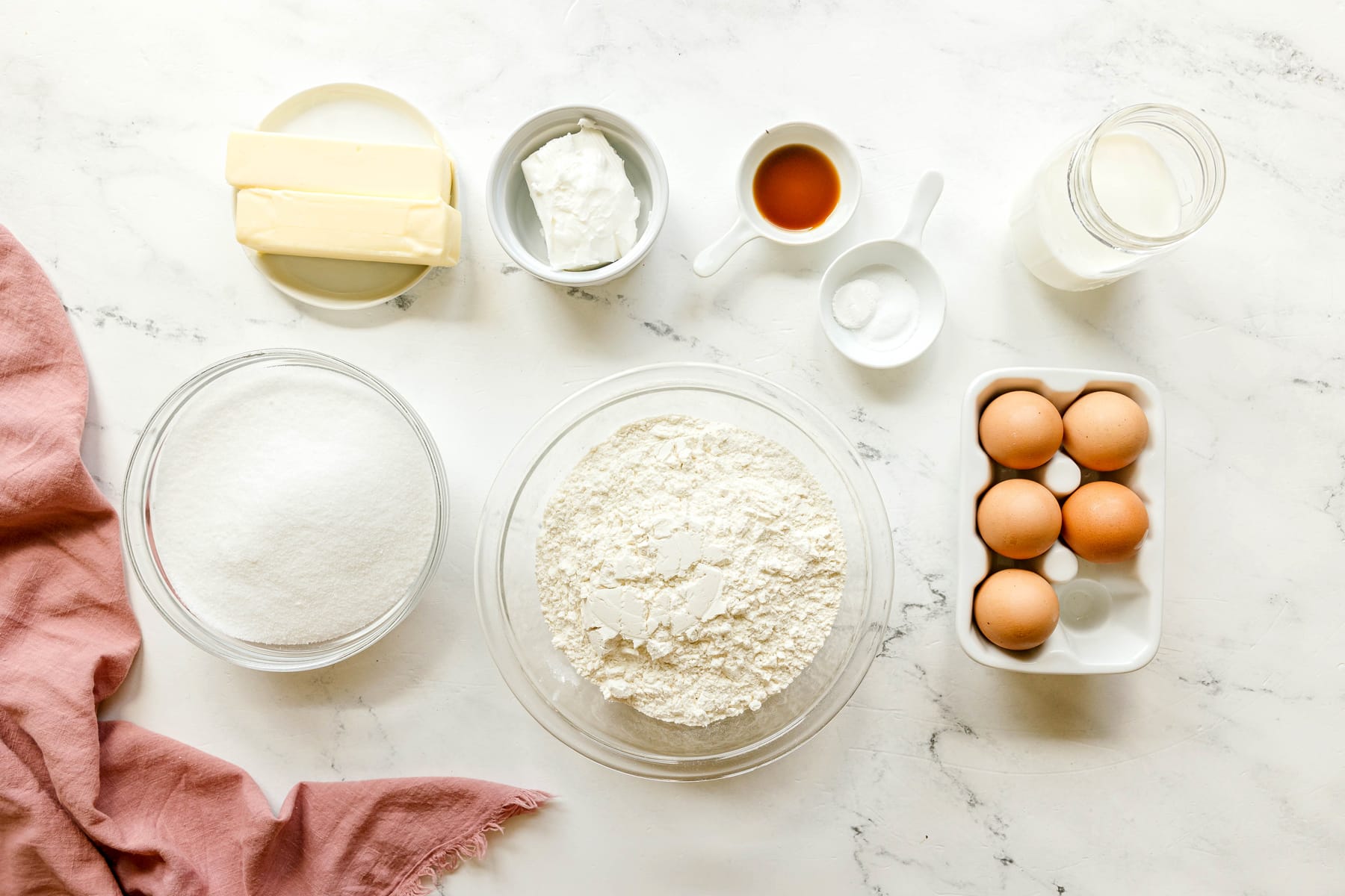 Baking ingredients for Paula Deen Pound Cake on a white surface, including unsalted butter, vegetable shortening, granulated sugar, all-purpose flour, baking powder, salt, large eggs, milk and vanilla extract, arranged neatly with a pink cloth on the side.