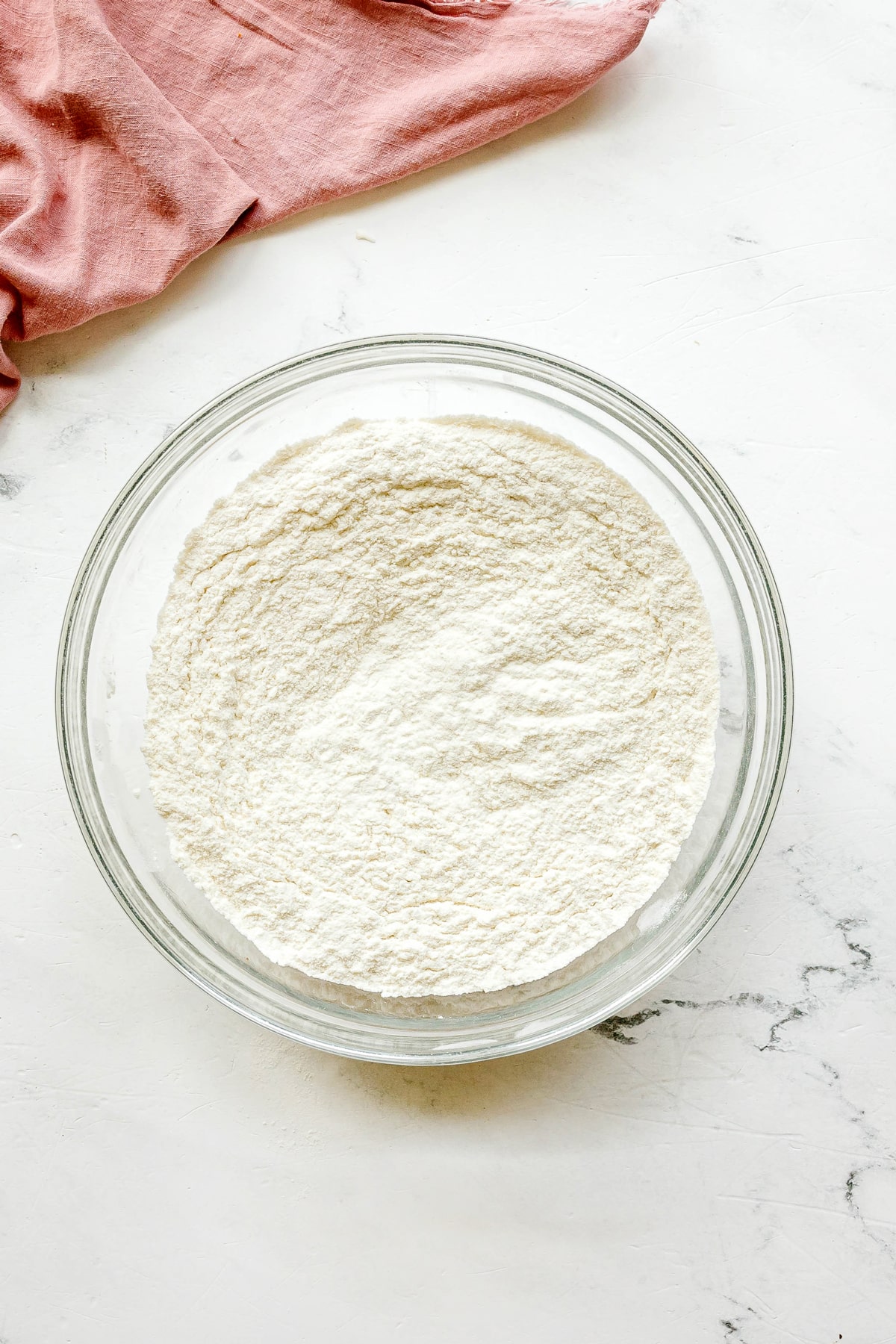 First step in preparing Paula Deen Pound Cake is to use a glass bowl filled and fill it with flour, baking powder and salt., on a white surface next to a pink cloth.