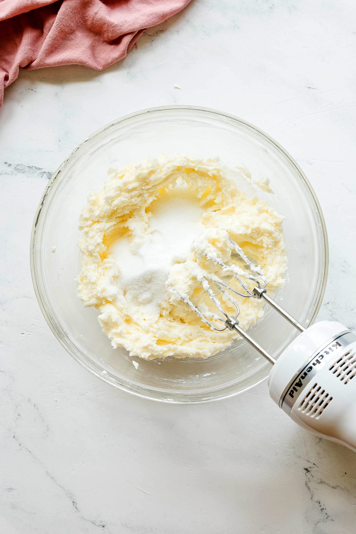 A glass bowl with softened butter and shortener is being mixed with an electric hand mixer on a white countertop. This is one step in preparing Paula Deen Pound Cake.