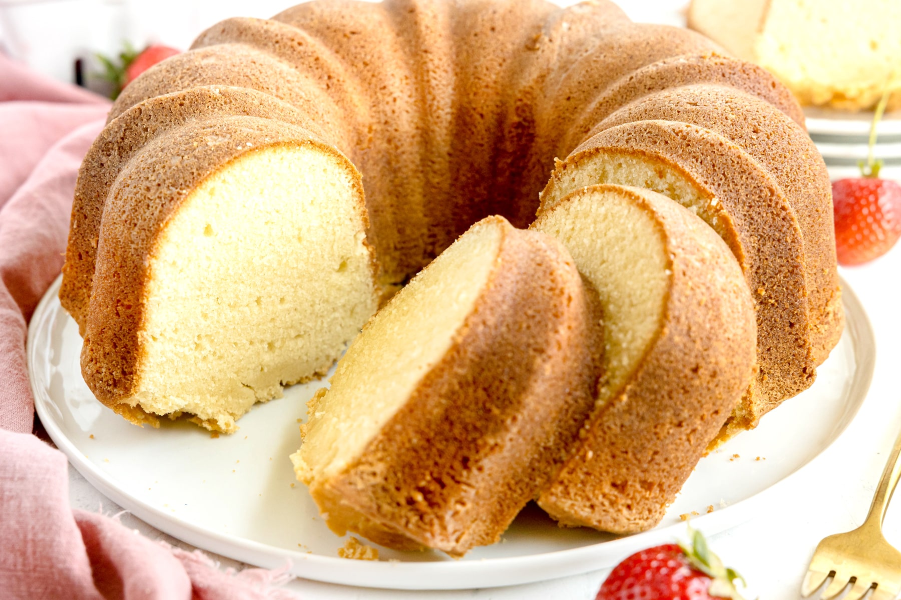 A whole Paula Deen Pound Cake with slices on a white plate. Linens, strawberries and utensils can also be seen.