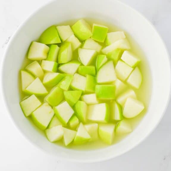 A white bowl filled with chopped green apple pieces soaking in water, viewed from above on a white surface.