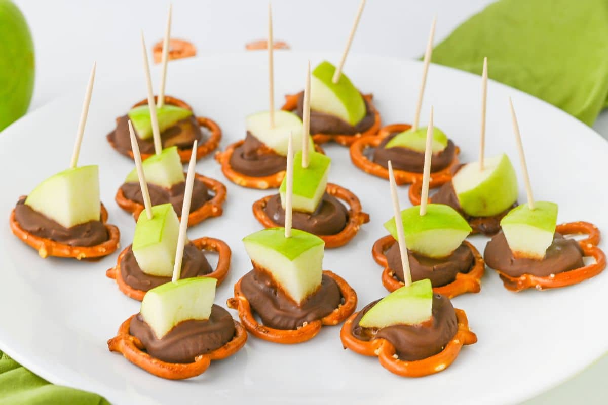 Green apple slices and chocolate on pretzels, each secured with a toothpick, arranged on a white plate.