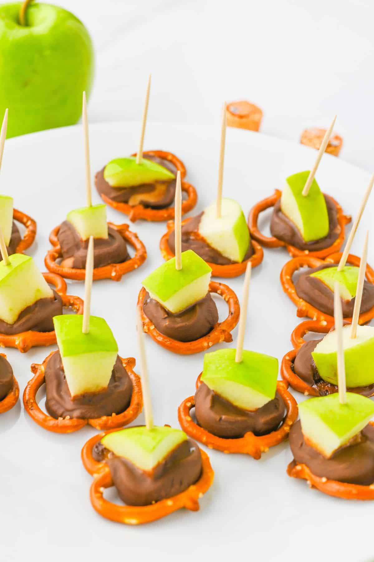 A plate of snacks with pretzels topped with chocolate and green apple chunks, each held together with a toothpick.