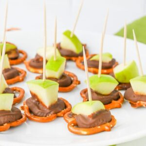 A white plate with snacks made of pretzels, chocolate, and green apple slices, each held together with a toothpick.