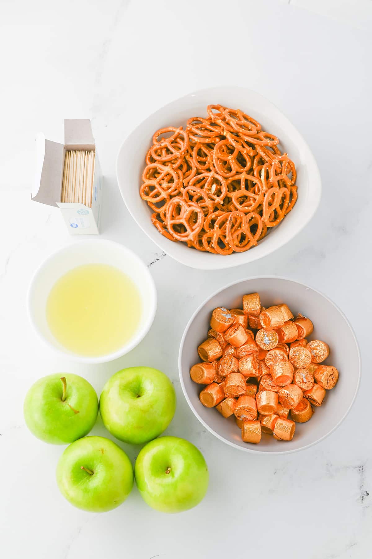 Three green apples, a bowl of mini pretzels, a bowl of caramel candies, a bowl of liquid, and a box of toothpicks are arranged on a white surface.