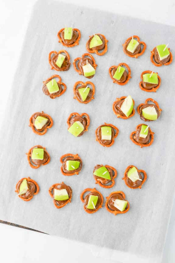 A baking sheet lined with parchment paper holding pretzel bites topped with melted chocolate and green apple pieces, some secured with toothpicks.