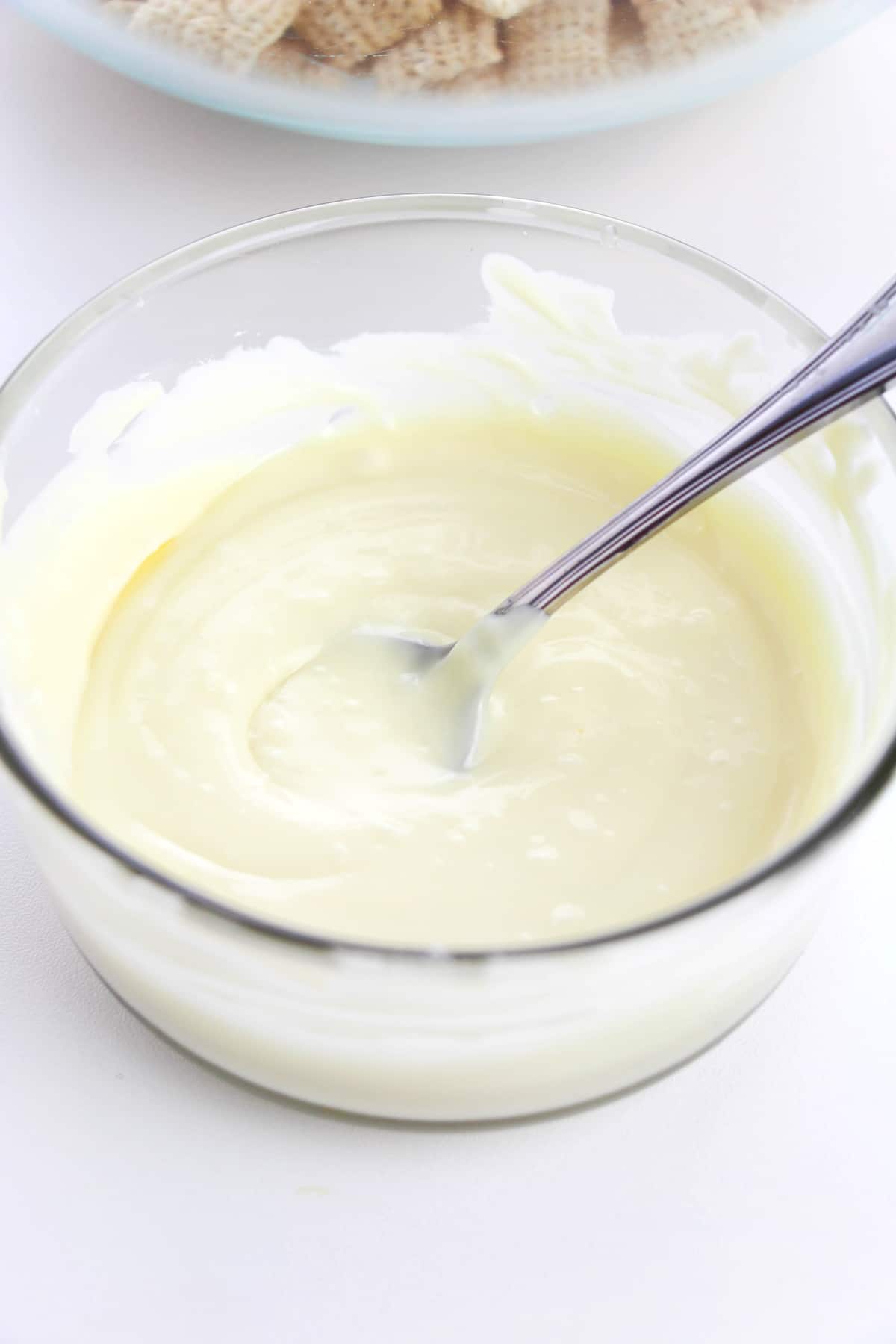 A clear glass bowl filled with melted white chocolate or cream mixture for Churro Muddy Buddies recipe , with a metal spoon resting inside.