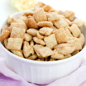 A white bowl filled with Churro Muddy Buddies recipe sits on a light pink cloth.