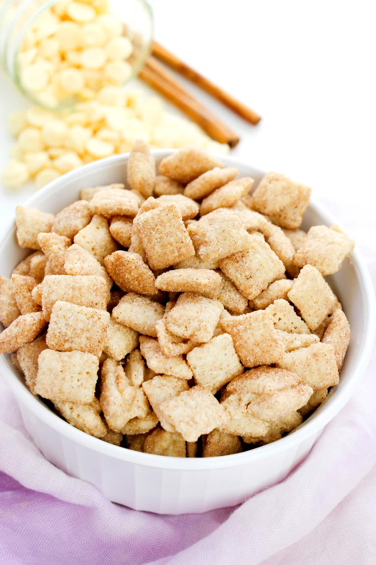 A white bowl filled with Churro Muddy Buddies recipe, with white chocolate chips and cinnamon sticks in the background.