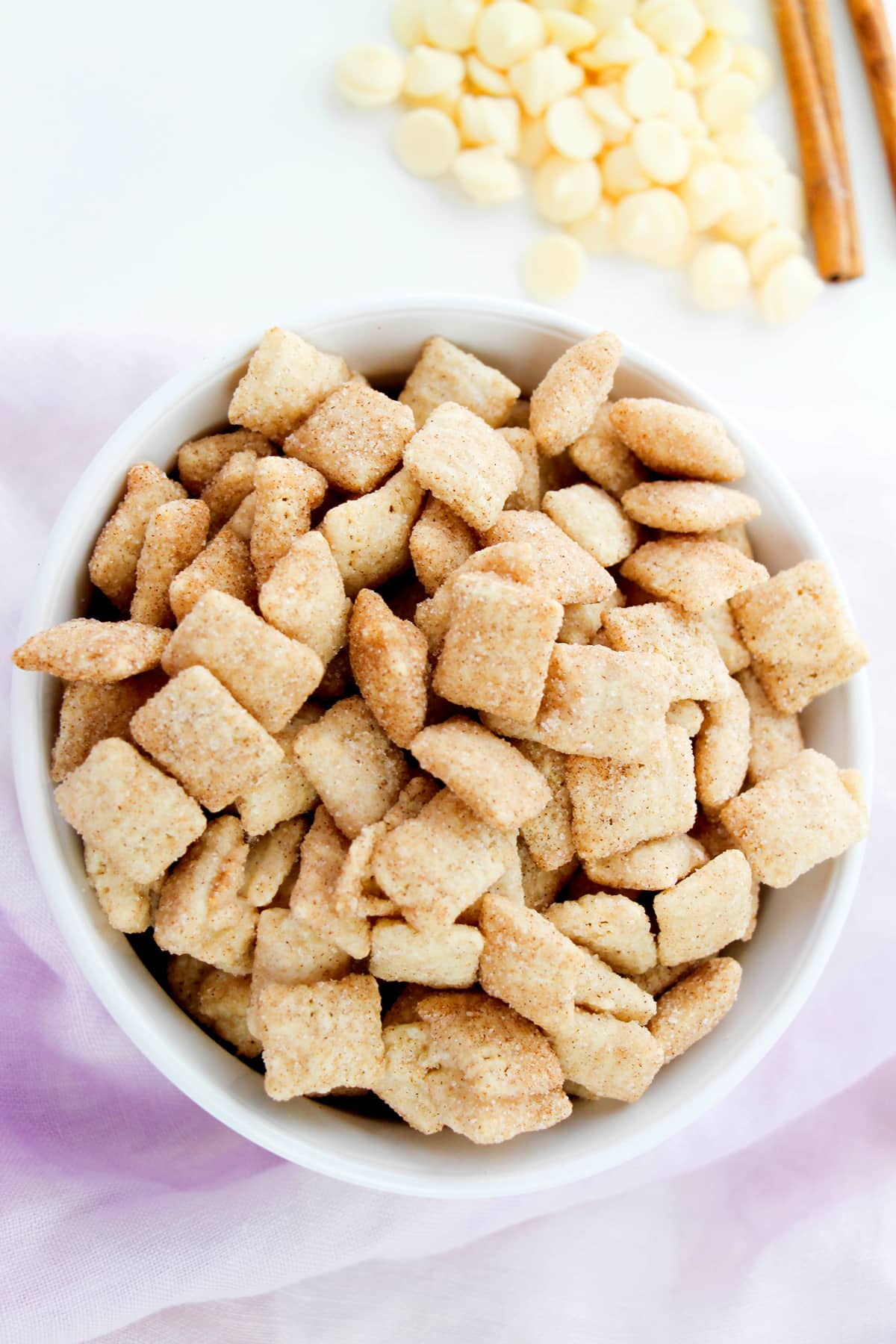 A white bowl filled with Churro Muddy Buddies recipe sits on a light pink cloth, with white chocolate chips and cinnamon sticks in the background.