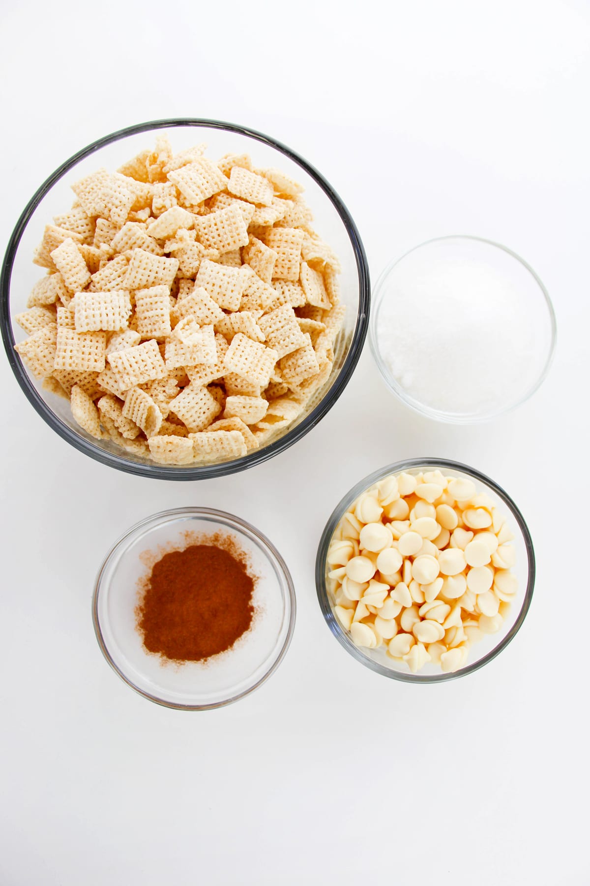 Four bowls with ingredients for Churro Muddy Buddies recipe: Chex cerea, white chocolate chips, white chocolate wafers, white sugar and cinnamon arranged on a white surface.
