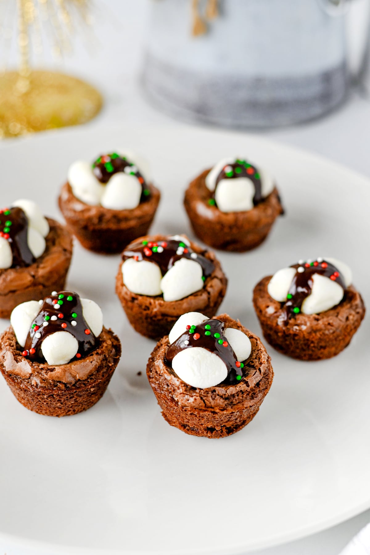 Six Hot Cocoa Brownies topped with marshmallows, chocolate sauce, and red and green sprinkles on a white plate.