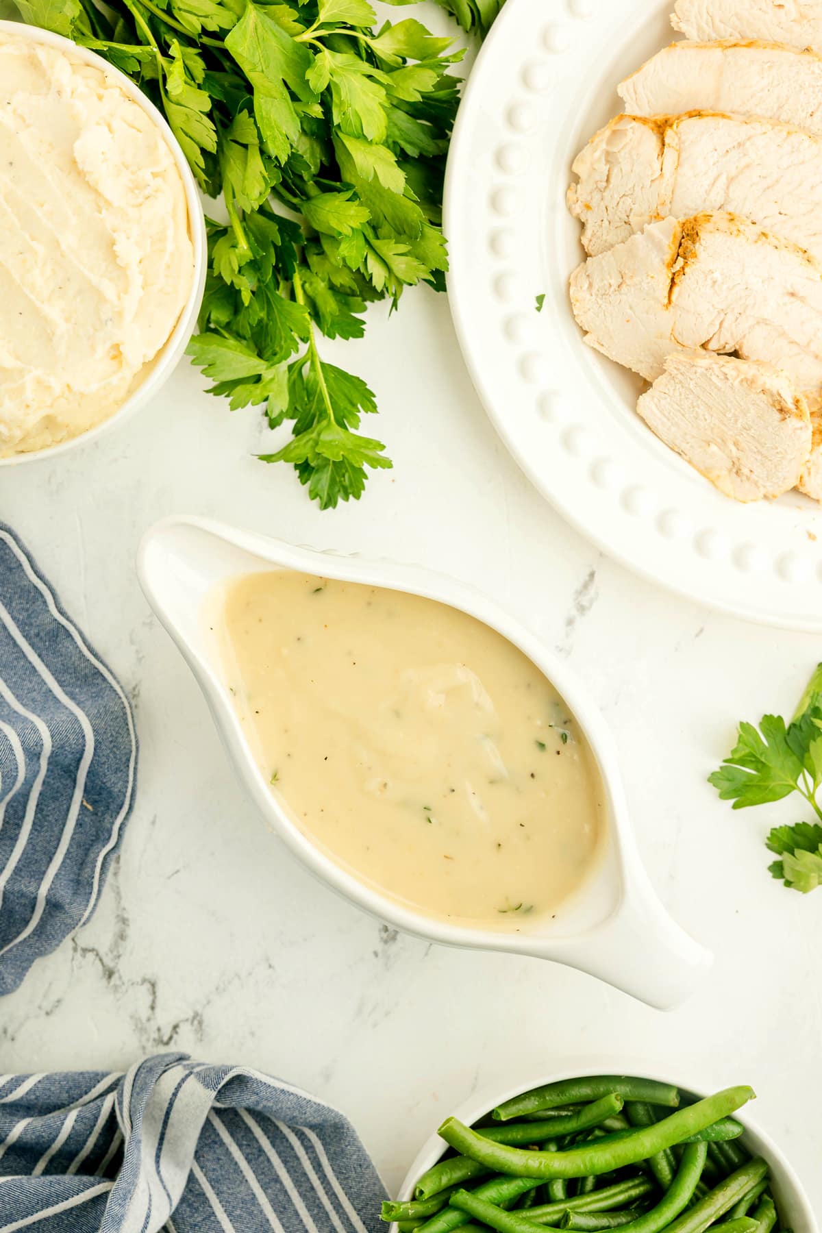A gravy boat filled with Chicken Gravy, surrounded by sliced chicken breast, mashed potatoes, and green beans.