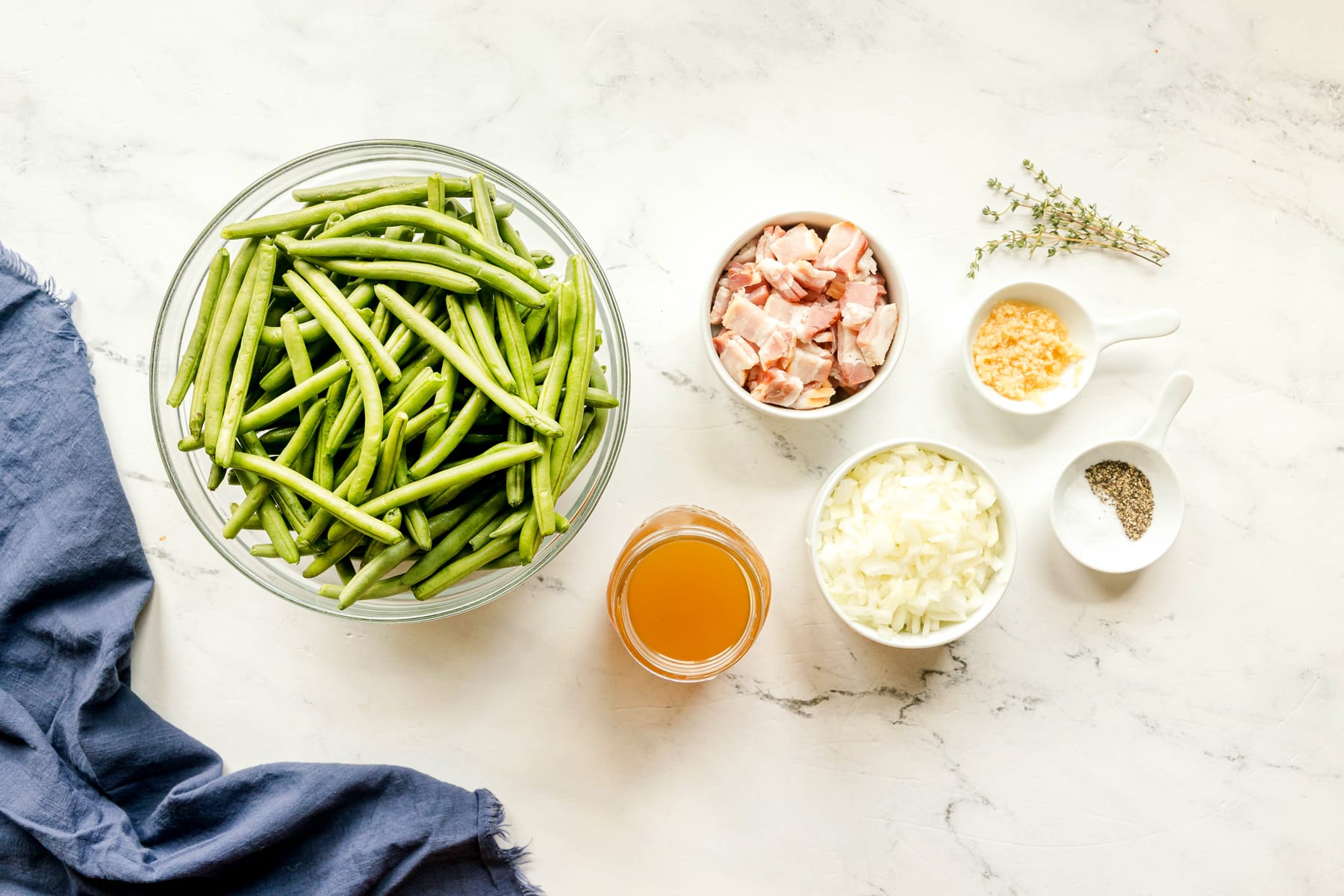 Ingredients for Southern Green Beans recipe are the following: green beans, thick-cut bacon, onion, garlic, chicken stock, fresh thyme, salt and black pepper on a white counter.