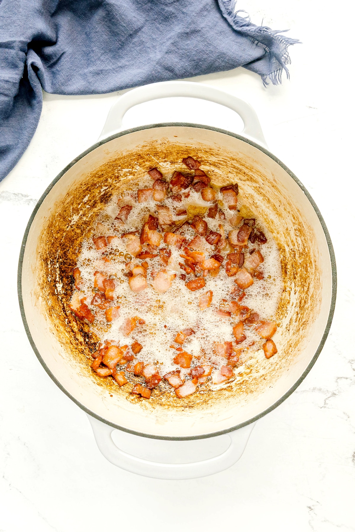 Bacon pieces sizzling in a large white enameled cast iron pot for Southern Green Beans recipe.