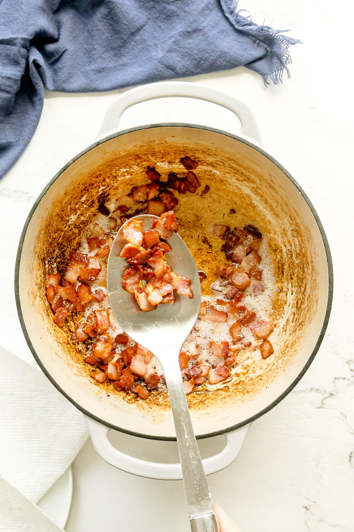 A spoonful of diced, fried bacon held over a Dutch oven containing more bacon pieces and rendered fat for Southern Green Beans recipe.