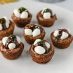Seven hot cocoa brownies topped with marshmallows, chocolate drizzle, and red and green sprinkles on a white plate.