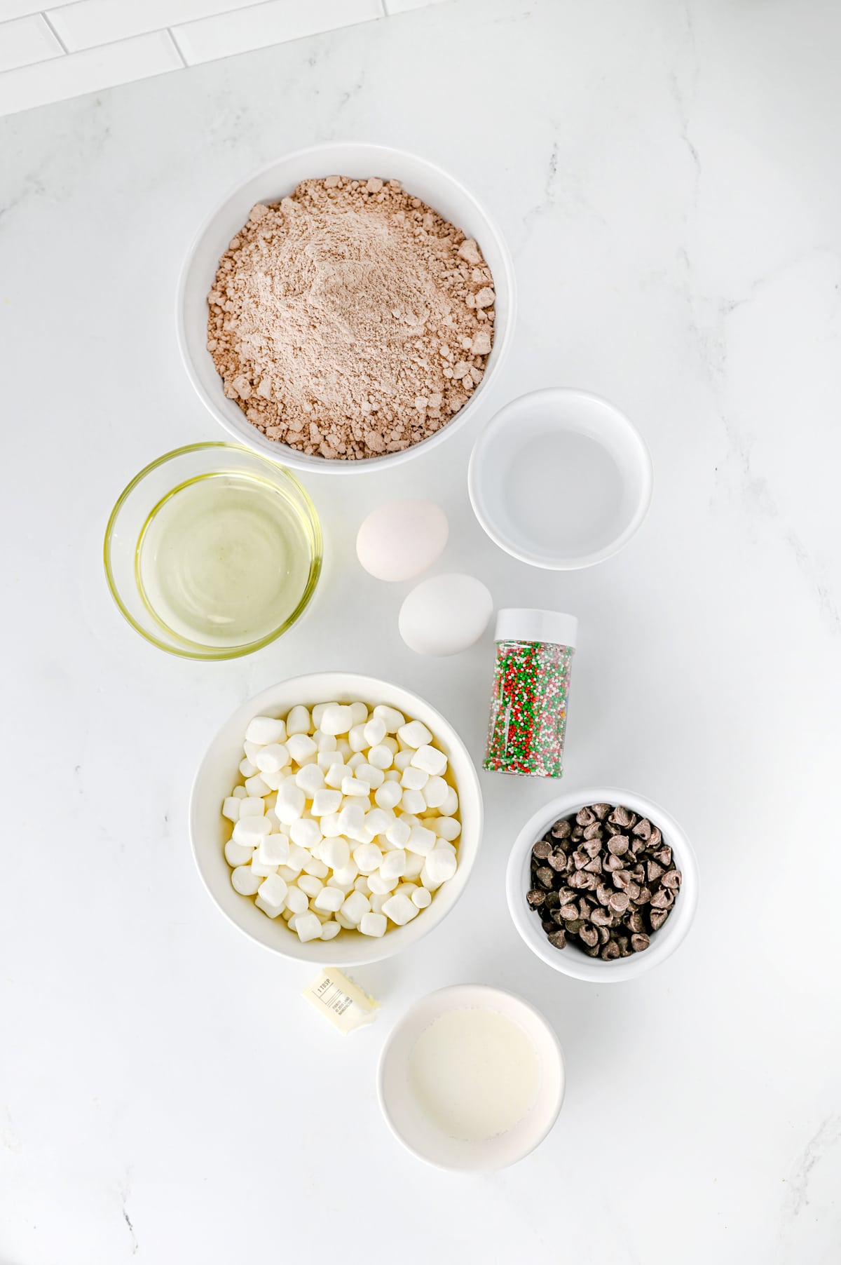 Baking ingredients for Hot Cocoa Brownies include brownie mix, water, oil, eggs, mini marshmallows, chocolate chips, Christmas sprinkles, and unsalted butter arranged on a marble surface.