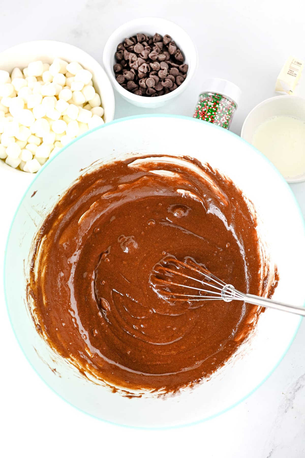 A whisk in a bowl of brownie mix surrounded by bowls of marshmallows, chocolate chips, sprinkles, and a small amount of liquid, for Hot Cocoa Brownies recipe.