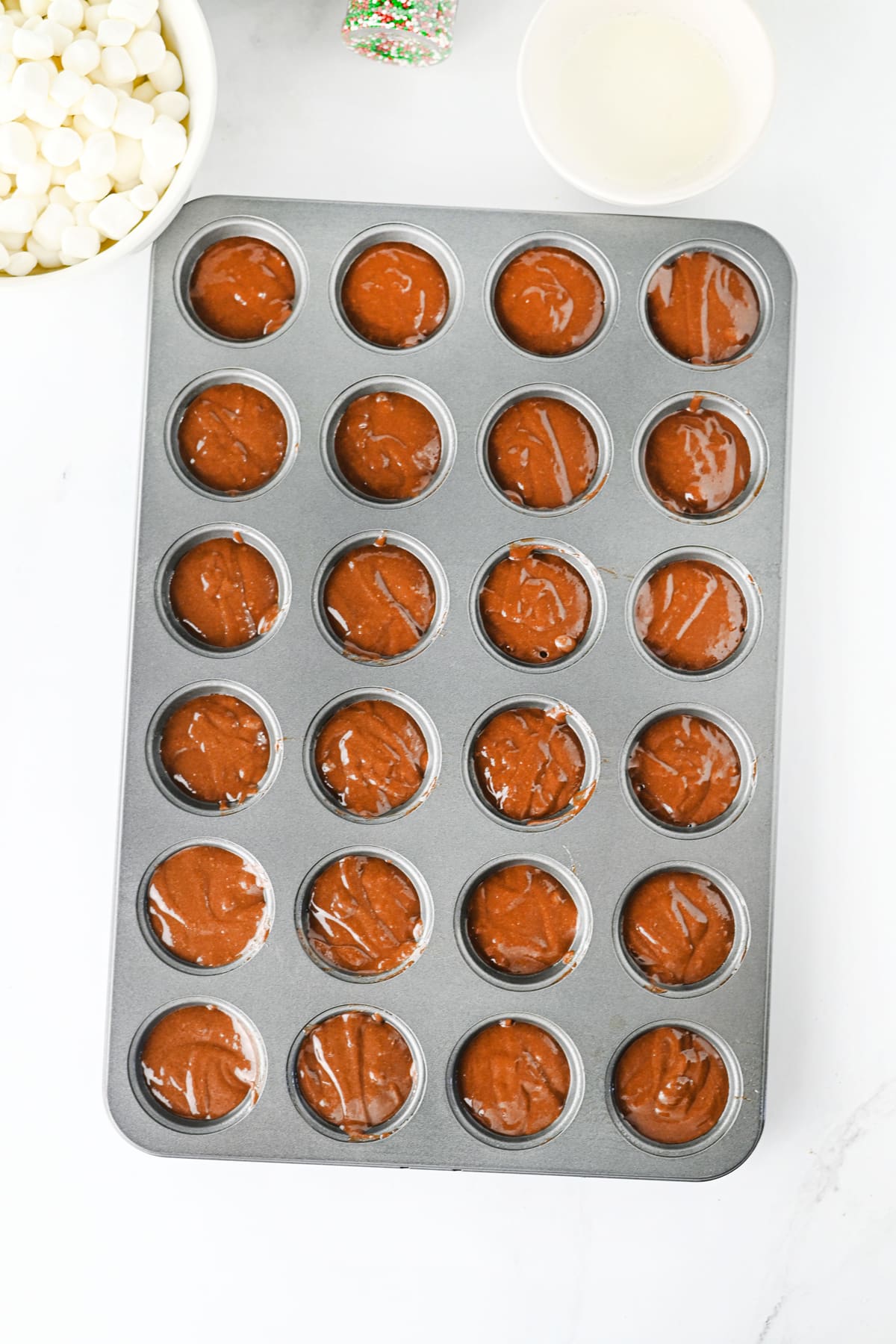 A 24-cup mini muffin pan filled with Hot Cocoa Brownies batter, with bowls of marshmallows and glaze nearby.