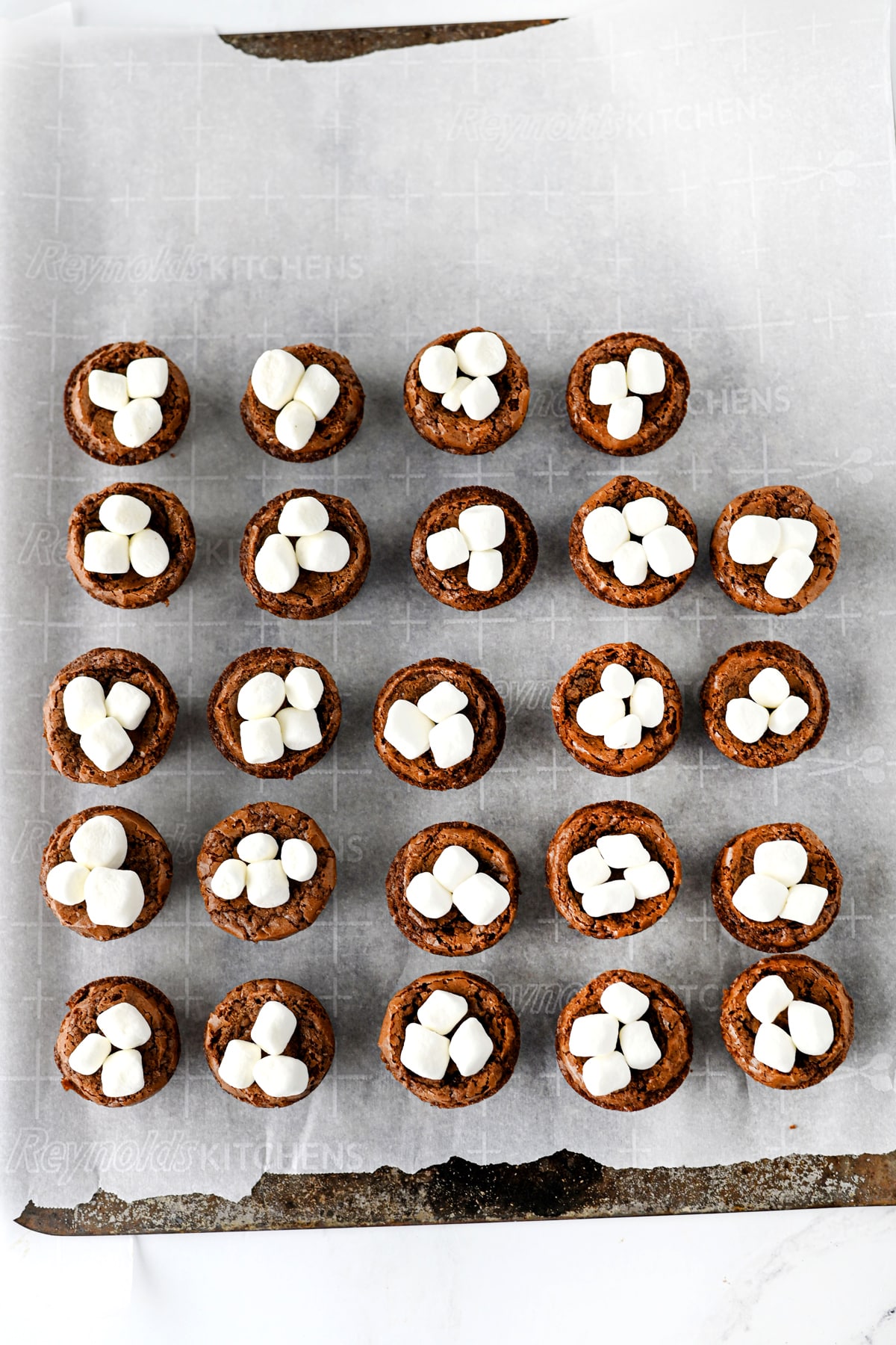 A grid of twenty-five round Hot Cocoa Brownies topped with mini marshmallows on parchment paper.