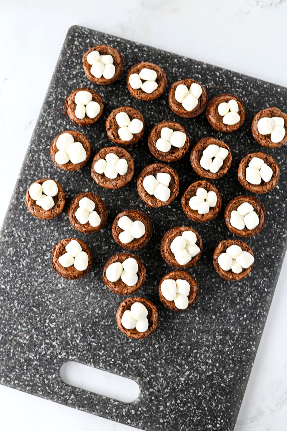 A collection of bite-sized Hot Cocoa Brownies topped with mini marshmallows arranged on a dark cutting board.
