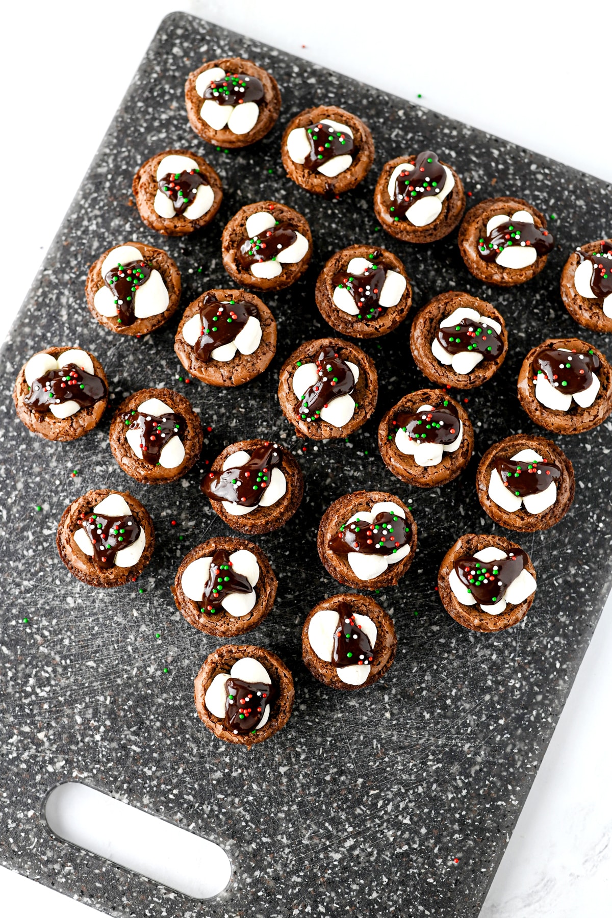 Hot Cocoa Brownies topped with marshmallows, chocolate drizzle, and holiday sprinkles on a dark speckled cutting board.